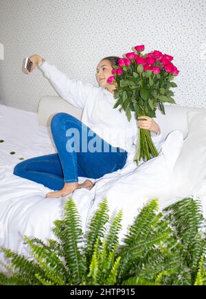 Ein junges Mädchen mit einem Strauß roter Rosen sitzt auf dem Bett und macht ein Selfie Stockfoto