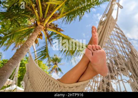 Entspannender Strandurlaub mit den Füßen der Frau in der Hängematte zwischen Kokospalme. Exotisches Resort auf tropischen Inseln. Sonniger, warmer Tag mit blauem Himmel. Stockfoto