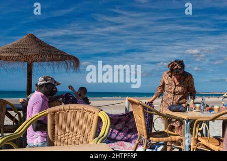 Afrikanischer Mann, der einer Frau am Strand in Conil de la Grenadine, Andalusien, Spanien, seine Waren zeigt Stockfoto