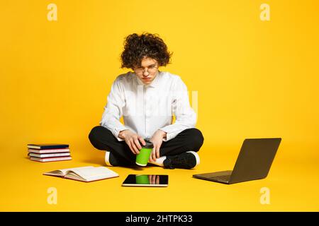 Porträt eines jungen Mannes, Studenten sitzen auf dem Boden, studieren, Vorbereitung für Prüfungen isoliert über gelben Studio-Hintergrund Stockfoto
