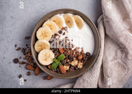 Draufsicht auf Müsli mit Joghurt, frischer Banane, Schokoladentropfen und Mandelnüssen für ein gesundes Frühstück auf grauem Beton-Hintergrund Stockfoto