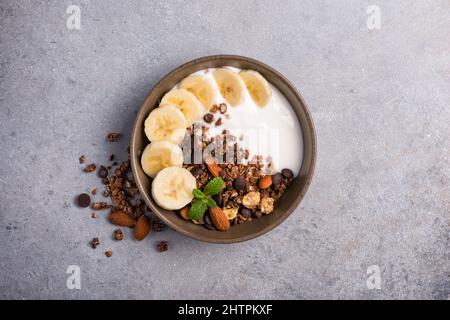 Draufsicht auf Müsli mit Joghurt, frischer Banane, Schokoladentropfen und Mandelnüssen für ein gesundes Frühstück auf grauem Beton-Hintergrund Stockfoto