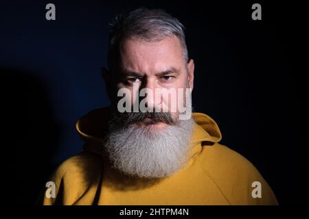Studioporträt eines reifen bärtigen Mannes mit gelbem Hoodie, der direkt auf die Kamera blickt, blauer Hintergrund Stockfoto