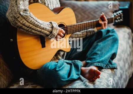 Junges Mädchen mit langen Nägeln spielt akustische Gitarre zu Hause. Teenager sitzt auf der Couch im Zimmer und lernt, Musikinstrumente zu spielen. Hintergrund. Nahaufnahme. Stockfoto