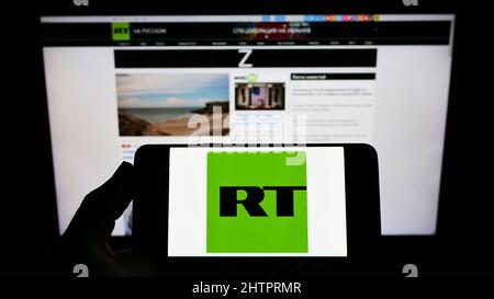 Person, die das Smartphone mit dem Logo des russischen staatlich kontrollierten Fernsehunternehmens RT auf dem Bildschirm vor der Website hält. Konzentrieren Sie sich auf die Telefonanzeige. Stockfoto