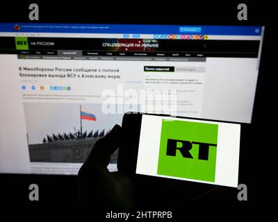 Person, die ein Mobiltelefon mit dem Logo des staatlichen russischen Fernsehunternehmens RT auf dem Bildschirm vor der Webseite hält. Konzentrieren Sie sich auf die Telefonanzeige. Stockfoto