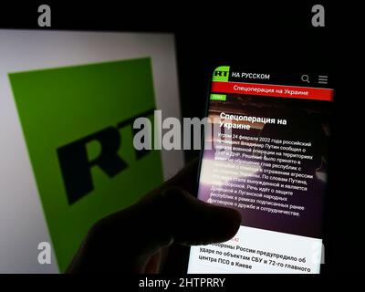 Person, die Mobiltelefon mit der Webseite des russischen staatlich kontrollierten Fernsehunternehmens RT auf dem Bildschirm mit Logo hält. Konzentrieren Sie sich auf die Mitte des Telefondisplays. Stockfoto