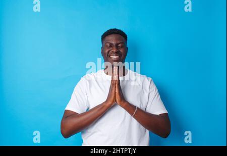 Ein Bild eines lächelnden Afrikaners in einem weißen T-Shirt, der seine Hände nahe seiner Brust auf einem isolierten blauen Hintergrund schubst. Stockfoto