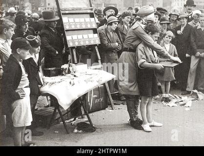 WHITBY-REGATTA um die 1920er Jahre - eine ungewöhnliche Wahrsagerin mit verbundenen Augen gibt einem Jungen eine Vorhersage, die ein Ticket an ihrem provisorischen Stand auf der jährlichen Regatta und Messe in Whitby, North Yorkshire, Großbritannien, gekauft hat. Die Mode der Zeit zeigt eine Vielzahl von Hüten und Mützen, die von Zuschauern getragen wurden, und dass junge Schuljungen immer noch kurze Hosen trugen. Stockfoto