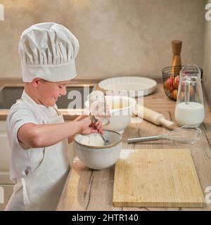 Ein Junge in der Kleidung des Küchenchefs gießt Mehl in den Teig, um Apfelkuchen zu kochen Stockfoto