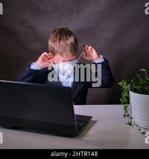 Ein Junge in einem Schulanzug mit einer medizinischen Maske auf seinem Gesicht an einem Computer, kopieren Raum auf einem dunklen Studiohintergrund Stockfoto