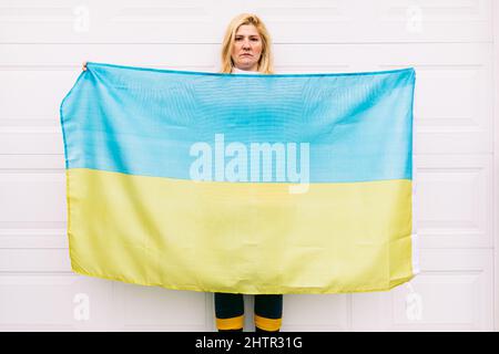 Ukrainische reife Frau mit blonden Haaren ernste Geste mit blauen und gelben ukrainischen Flagge auf weißem Hintergrund auf der Straße. Ukraine Krieg, Invasion, p Stockfoto