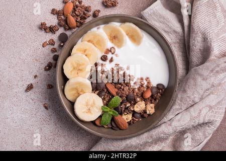 Draufsicht auf Müsli mit Joghurt, frischer Banane, Schokoladentropfen und Mandelnüssen für ein gesundes Frühstück auf braunem Betongrund Stockfoto