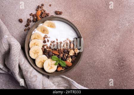 Draufsicht auf Müsli mit Joghurt, frischer Banane, Schokoladentropfen und Mandelnüssen für ein gesundes Frühstück auf braunem Betongrund Stockfoto