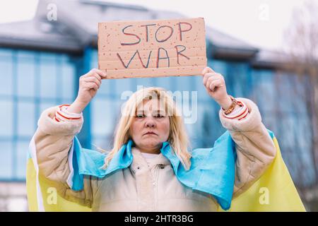 Ukrainische Frau mit blonden Haaren und ernster Geste, mit einer blau-gelben ukrainischen Flagge mit einem Schild, das sagt: "Stop war", auf der Straße, protestiert Stockfoto