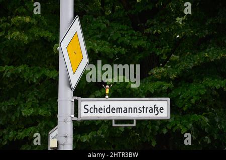 Berliner Straßenschild der Scheidemannstraße mit kleinem Korkmann darauf und einem Wegweiser mit grünen Bäumen im Hintergrund Stockfoto