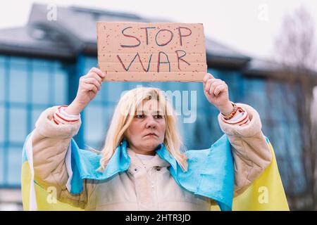 Ukrainische Frau mit blonden Haaren und ernster Geste, mit einer blau-gelben ukrainischen Flagge mit einem Schild, das sagt: "Stop war", auf der Straße, protestiert Stockfoto