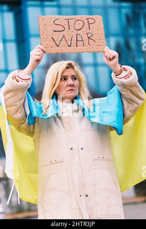 Ukrainische Frau mit blonden Haaren und ernster Geste, mit einer blau-gelben ukrainischen Flagge mit einem Schild, das sagt: "Stop war", auf der Straße, protestiert Stockfoto