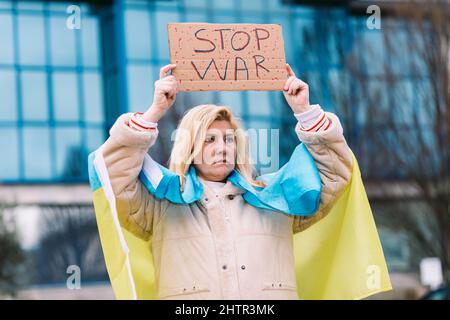 Ukrainische Frau mit blonden Haaren und ernster Geste, mit einer blau-gelben ukrainischen Flagge mit einem Schild, das sagt: "Stop war", auf der Straße, protestiert Stockfoto
