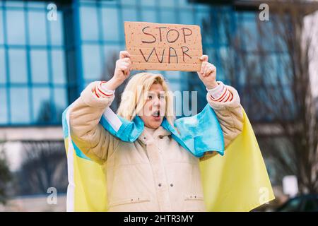 Ukrainische Frau mit blonden Haaren und ernster Geste, mit einer blau-gelben ukrainischen Flagge mit einem Schild, das sagt: "Stop war", auf der Straße, schreiend Stockfoto