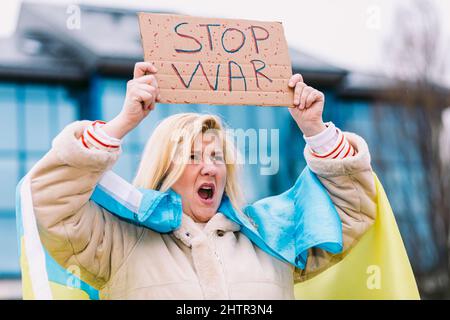 Ukrainische Frau mit blonden Haaren und ernster Geste, mit einer blau-gelben ukrainischen Flagge mit einem Schild, das sagt: "Stop war", auf der Straße, schreiend Stockfoto