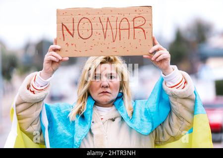 Ukrainische Frau mit blonden Haaren und ernster Geste, mit einer blau-gelben ukrainischen Flagge mit einem Schild, das sagt: "Kein Krieg", auf der Straße, protestierend Stockfoto