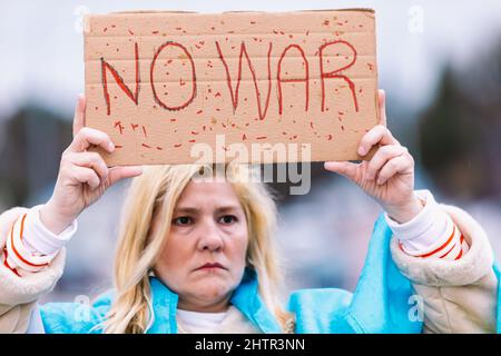 Ukrainische Frau mit blonden Haaren und ernster Geste, mit einer blau-gelben ukrainischen Flagge mit einem Schild, das sagt: "Kein Krieg", auf der Straße, protestierend Stockfoto