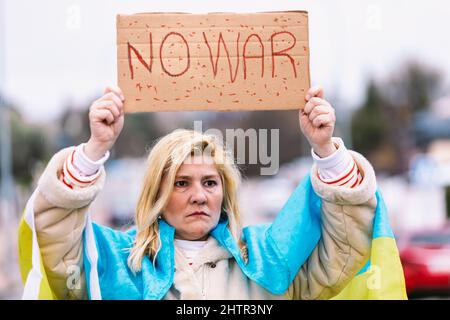 Ukrainische Frau mit blonden Haaren und ernster Geste, mit einer blau-gelben ukrainischen Flagge mit einem Schild, das sagt: "Kein Krieg", auf der Straße, protestierend Stockfoto