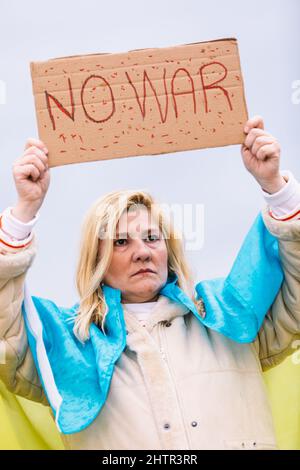 Ukrainische Frau mit blonden Haaren und ernster Geste, mit einer blau-gelben ukrainischen Flagge mit einem Schild, das sagt: "Kein Krieg", auf der Straße, protestierend Stockfoto