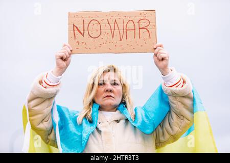 Ukrainische Frau mit blonden Haaren und ernster Geste, mit einer blau-gelben ukrainischen Flagge mit einem Schild, das sagt: "Kein Krieg", auf der Straße, protestierend Stockfoto