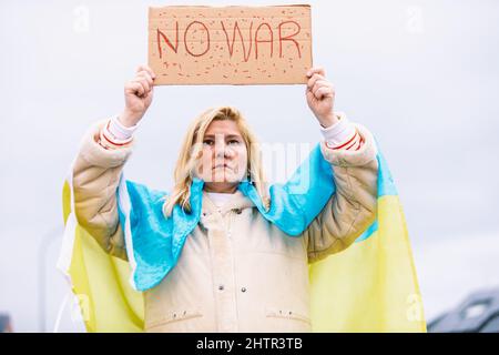Ukrainische Frau mit blonden Haaren und ernster Geste, mit einer blau-gelben ukrainischen Flagge mit einem Schild, das sagt: "Kein Krieg", auf der Straße, protestierend Stockfoto