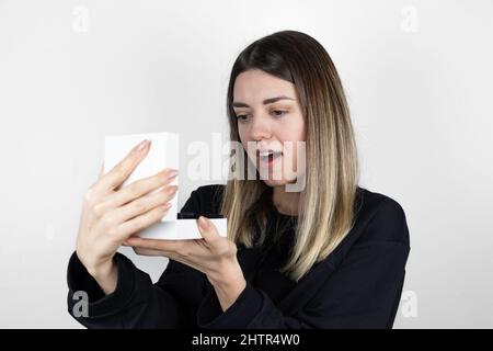 Mädchen hält eine kleine weiße Schachtel in der Hand. Als sie das Geschenk sieht, ist sie glücklich und überrascht. „Was haben wir hier?“ Konzeptfoto in Weiß. Stockfoto