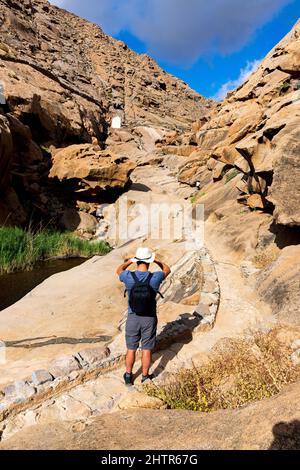 Wanderer mit Rucksack mit Blick auf die alte Kapelle zwischen Felsschluchten, Barranco de las Penitas, Fuerteventura, Kanarische Inseln, Spanien Stockfoto