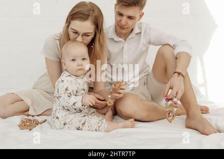 Porträt einer jungen Familie mit einem Cherubim-Kleinkind, mit blauen Augen, die einen natürlichen Beißring aus Holz auf einem weißen Bett halten. Stockfoto