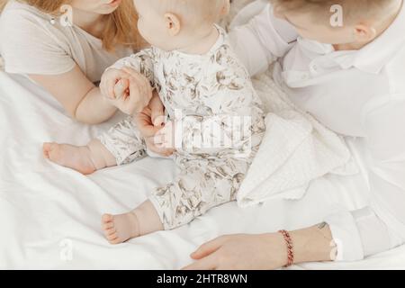 Draufsicht auf junge Familie in weißen Kleidern auf weißen Baumwollbettwäsche mit niedlichen kleinen blauen Augen Baby sitzen. Stockfoto