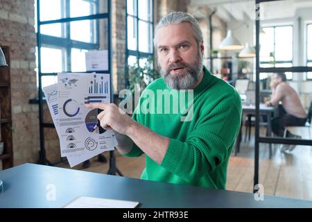 Porträt eines bärtigen Geschäftsmannes, der auf den Finanzbericht zeigt und die darauf abdruckende Grafik während der Sitzung im Vorstandszimmer erklärt Stockfoto