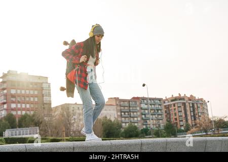 Junge Latina, die mit Rucksack und Skateboard durch die Stadt läuft. Venezolanische hispanische Frau, die Sport praktiziert. Stockfoto