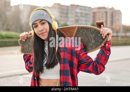 Porträt einer jungen lächelnden Skateboarderin, die ihr Skateboard hält. Lateinische Frau, die im Freien auf die Kamera schaut. Stockfoto