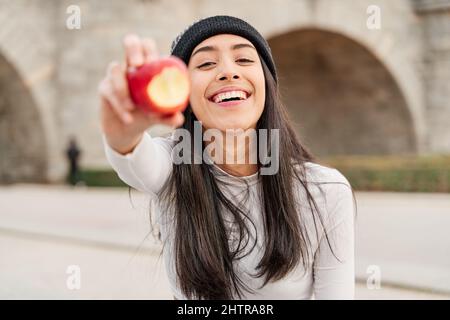 Junge und schöne lateinische Frau hält einen gebissenen Apfel und zeigt ihn der Kamera. Frau mit perfekten Zähnen essen gesund, gesundes Leben Konzept Stockfoto
