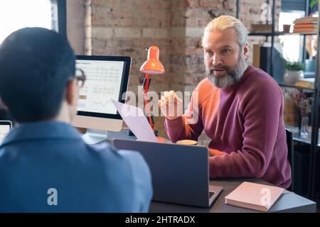 Ein reifer bärtiger Geschäftsmann hält ein Dokument und spricht mit seinen Kollegen bei der Besprechung über die geschäftliche Innovation Stockfoto