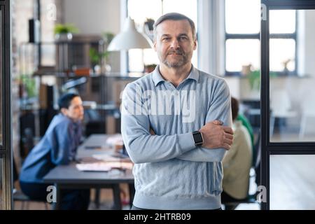 Porträt eines reifen, selbstbewussten Geschäftsmannes in blauem Hemd, der mit gekreuzten Armen steht und während eines Geschäftstreffens im Hintergrund auf die Kamera blickt Stockfoto