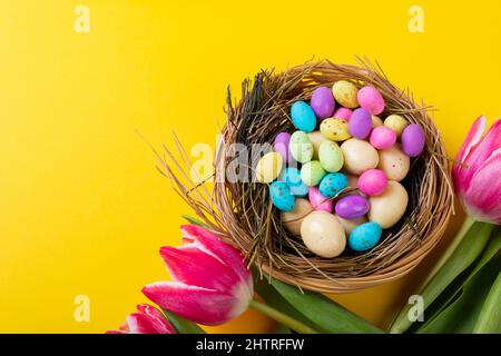 Nahaufnahme von bunten ostereiern und Süßigkeiten im Nest mit Tulpen auf gelbem Hintergrund Stockfoto