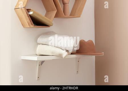 Regal mit einem Stapel warmer Pullover und einem Hut, der an einer hellen Wand hängt Stockfoto