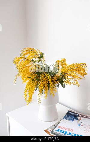 Vase mit Mimosenblüten und Zeitschriften auf Kommode in der Nähe einer hellen Wand Stockfoto