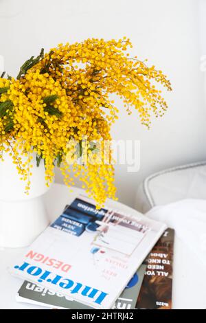 Vase mit Mimosenblumen und Zeitschriften auf Nachttisch in der Nähe der hellen Wand, Nahaufnahme Stockfoto