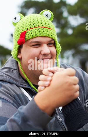 Spielerische Rivalität. Ein junger Mann, der eine froschförmige Mütze trägt, beschäftigt sich mit Armringen draußen. Stockfoto