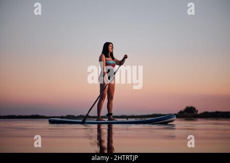 SUP-Boarding Frau mittleren Alters rudert mit Pfütze, die auf den welligen See blickt, mit einem erstaunlichen rosa Himmel im Hintergrund, der am Abend Badeanzug trägt Stockfoto