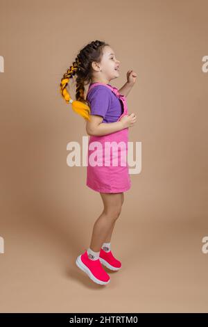 Energisches weibliches Kind im Springen mit zwei gelben Kanekalon-Zöpfen, die einen pinken Jumpsuit, ein violettes T-Shirt und rosa Sneakers tragen Stockfoto