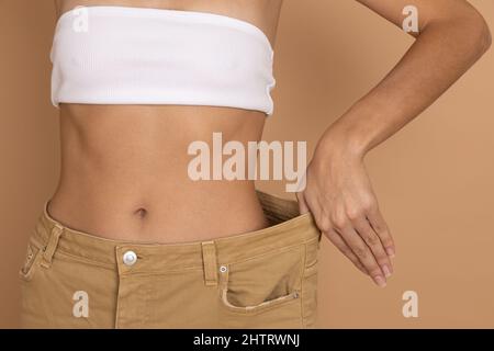 Nahaufnahme der Frau Taille in großen übergroßen Jeans mit Hand zeigt gute schlanke Form auf körperfarbenen Hintergrund. Gewichtsverlust durch die Aufrechterhaltung der Ernährung und Bewegung Stockfoto