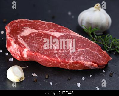 RAW Steak Ribeye mit Knoblauch und Gewürzen auf schwarzem Schieferstein Hintergrund, Seitenansicht eines marmorierten, frischen Rindersteaks Stockfoto